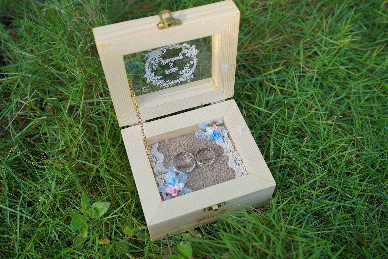 gallery-1 A rustic wooden wedding ring box with engraved glass and wedding bands, placed on grass.