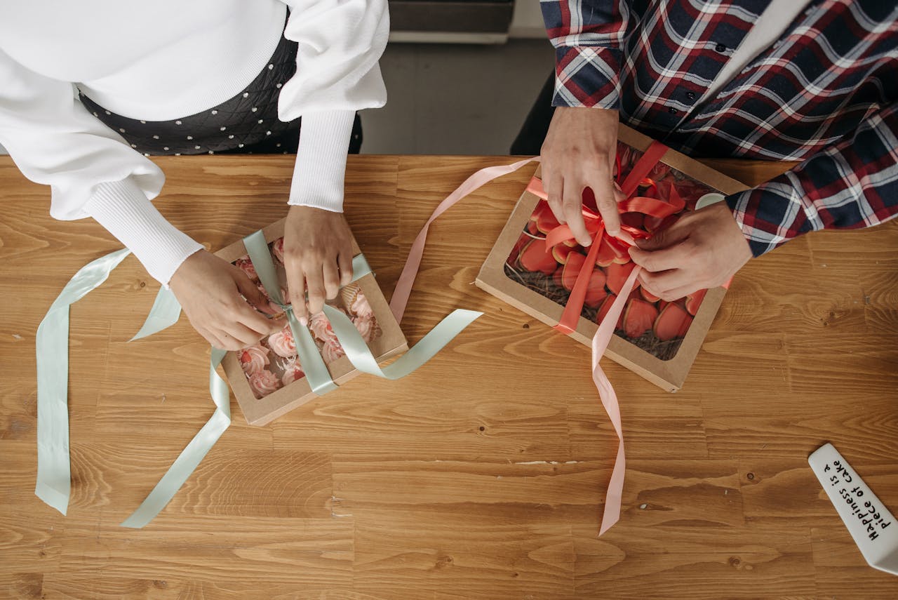 services-01 Overhead view of a couple wrapping gift boxes with pink and turquoise ribbons on a wooden table.