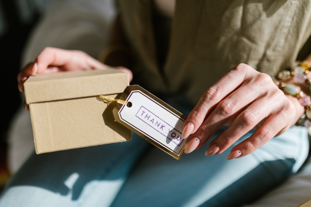 hero-img-01 Close-up of hands holding a gift box with a Thank You tag attached, in natural lighting.